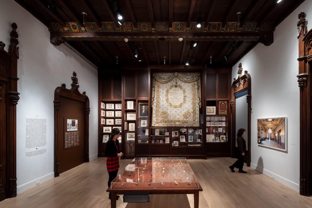 Museum gallery with wood-paneled display cases featuring documents, photographs, and a large embroidered textile; two visitors are present.