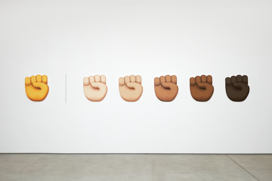 Six large fist emojis mounted on a white wall, progressing from yellow on the left to a range of skin tones from light to dark on the right, separated by a vertical line between the yellow fist and the others.