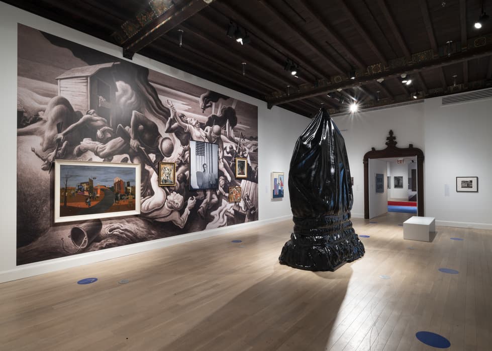 Gallery space with large black-and-white mural of writhing figures on the back wall, several framed artworks mounted on top of it; in the foreground stands a tall, black, glossy, shrouded sculpture resembling a hooded figure, on light wood floors under a dark wooden ceiling.