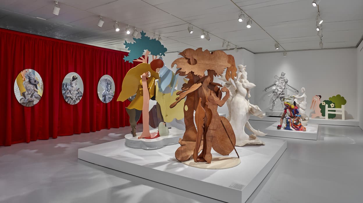 Gallery view of Rachel Feinstein exhibition with colorful, multi-material figurative sculptures on white platforms, red curtains with framed oval portraits, and dramatic overhead lighting.