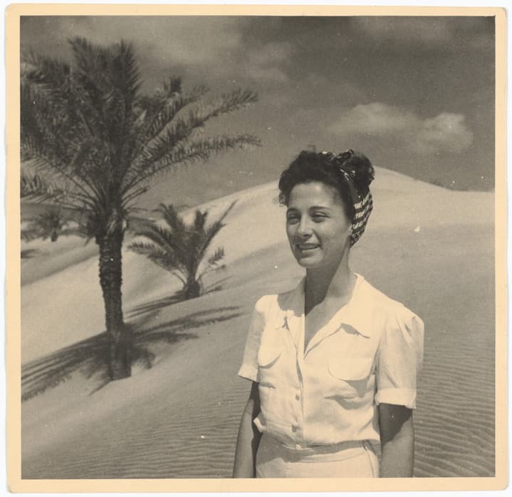 Black-and-white photograph of a woman in a short-sleeve blouse and headscarf standing in a desert landscape with sand dunes and palm trees under a partly cloudy sky.