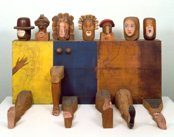Colorful wooden sculpture featuring a rectangular base divided into yellow, blue, and brown panels. Seven carved wooden heads with distinct facial expressions and hairstyles are mounted on top of the base. Various carved wooden limbs—arms and legs—are attached to the front and sides, giving the piece a playful, anthropomorphic appearance. The overall composition combines abstract geometry with figural elements.