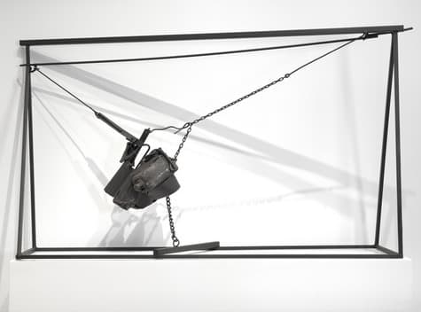 Metal sculpture composed of a rectangular black steel frame supporting a suspended arrangement of industrial-looking metal objects. Chains and rods hold a central cluster of dark, roughly textured metal parts, with geometric tension created by the angled supports and suspended elements. The structure casts sharp shadows on the white wall behind it.