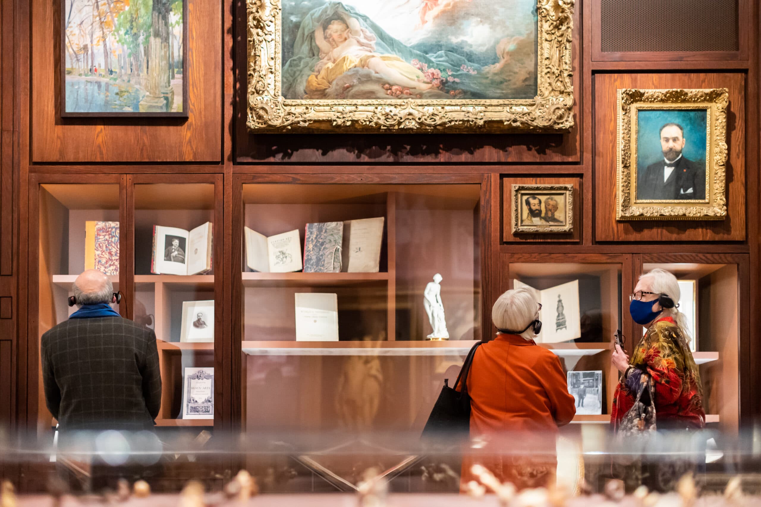 Visitors viewing a wood-paneled museum display with books, small sculptures, and framed artworks.