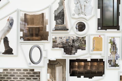 Wall installation of various framed photographs and objects, including architectural fragments, a white bird, empty theater seats, and abstract images, arranged in an irregular collage-like composition.