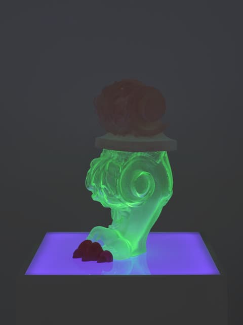 Contemporary sculpture featuring a glowing green, ornate column fragment topped with a reddish organic form, displayed on a pedestal lit with purple light in a dark gallery space.