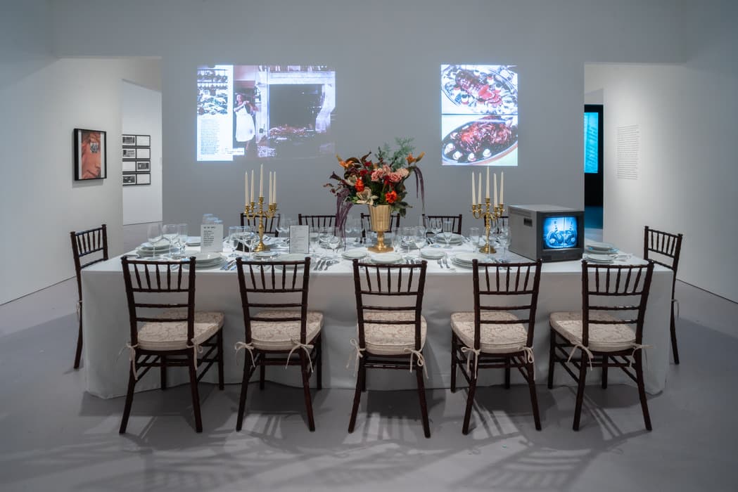 Art installation of a long dining table set with formal place settings, floral centerpiece, and golden candelabras, surrounded by wooden chairs with cream cushions. Behind the table, two food-related images are projected on the wall, and a small CRT monitor displaying a food scene is placed on the table's right end. The gallery setting includes other framed artworks on the walls.